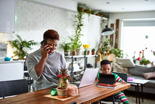 A woman on the phone works on a laptop at a wooden table, while a child in pajamas uses a tablet.