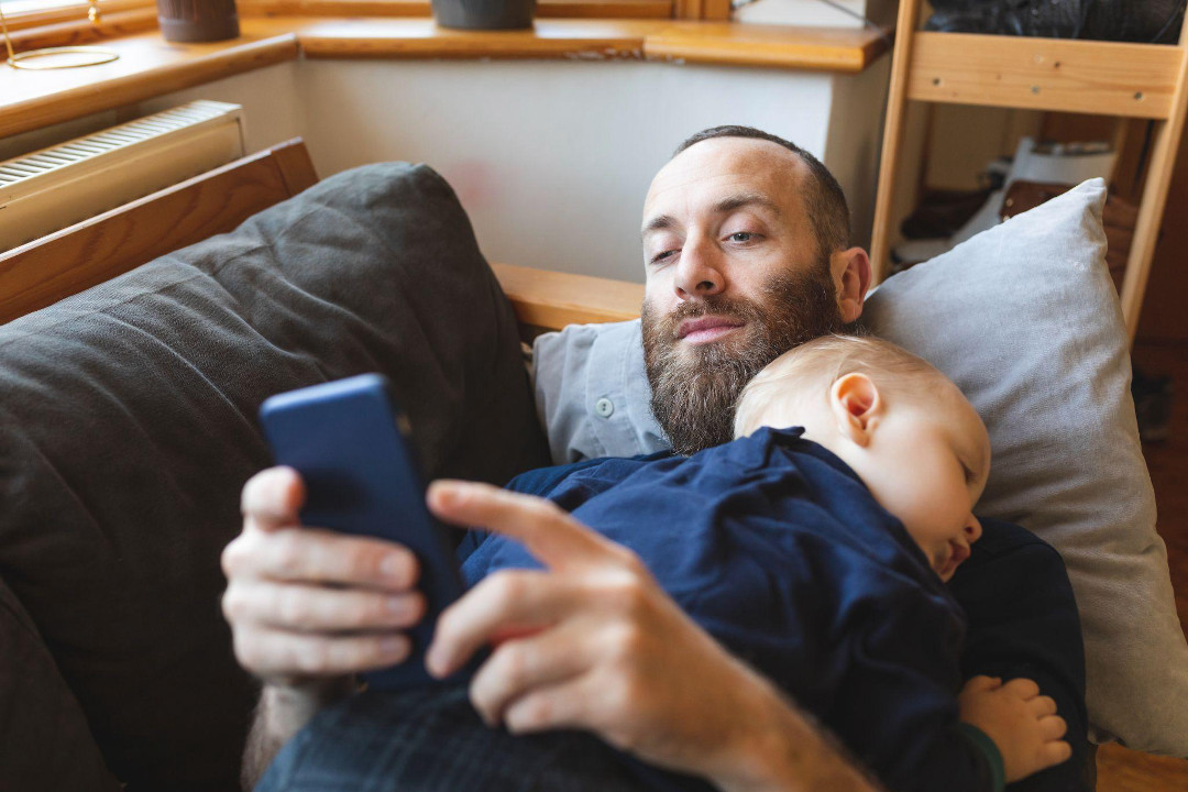 A bearded man lies on a couch with a sleeping toddler on his chest, holding a smartphone.