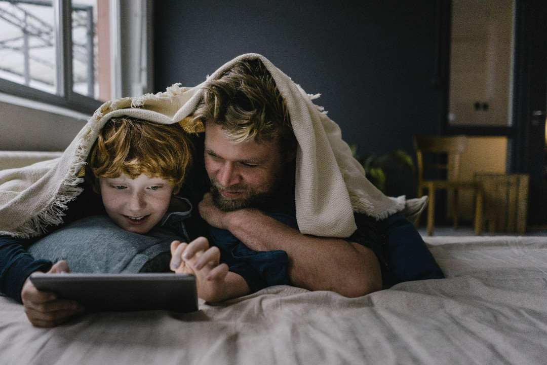 Father and son under a blanket, smiling and using a tablet in a cozy room.