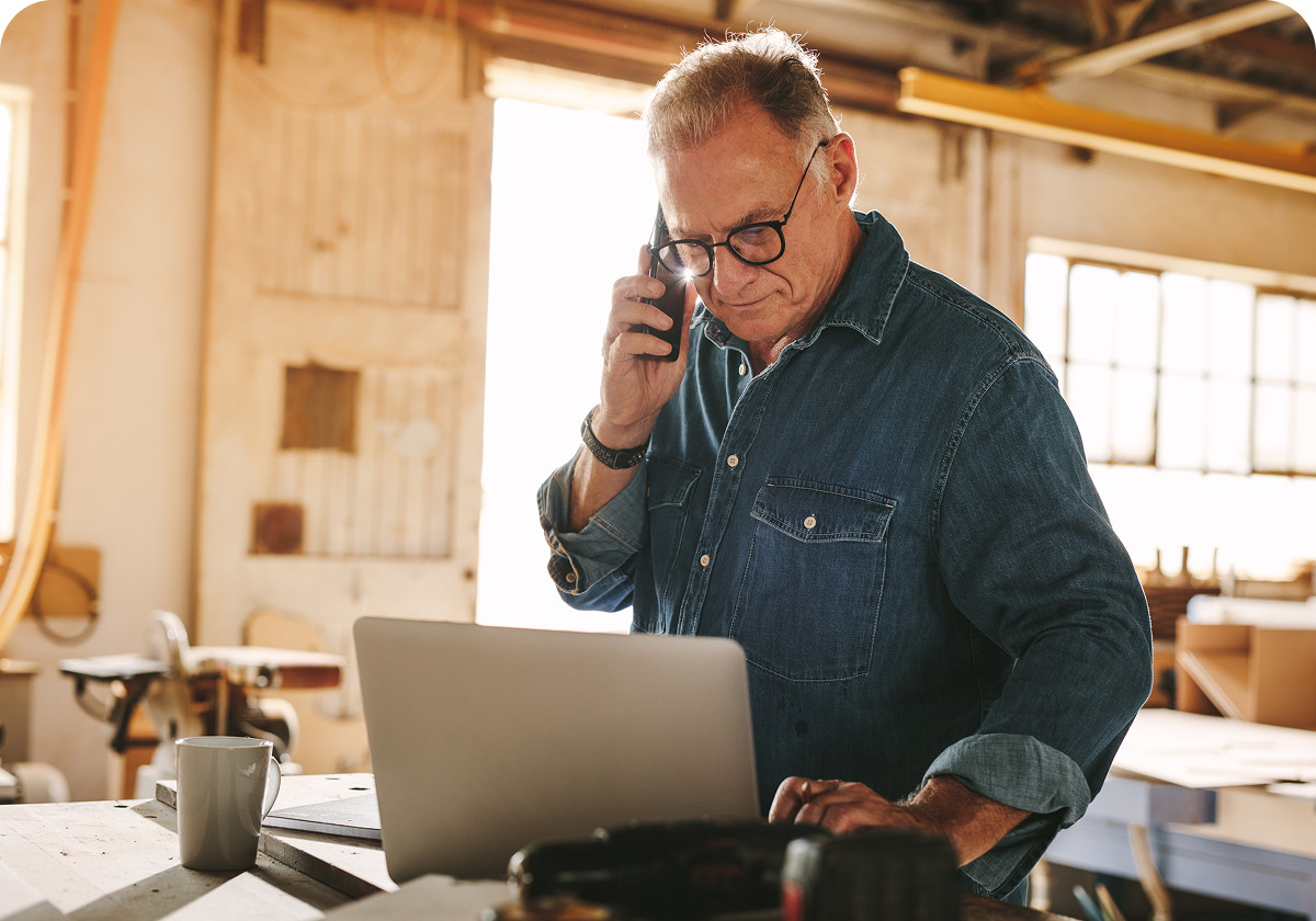Middle-aged man in denim shirt talks on phone, works on laptop in a woodworking shop.