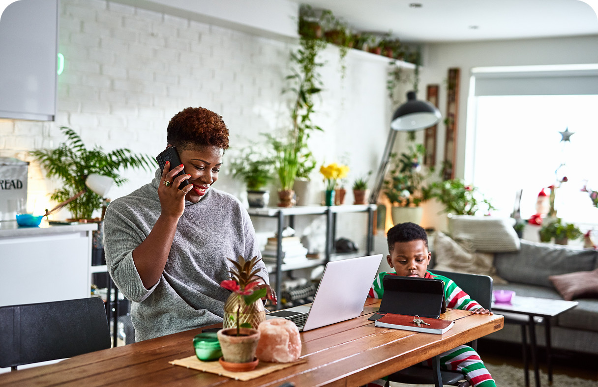 A woman works on a laptop and talks on the phone at a table, while a child in pajamas uses a tablet.