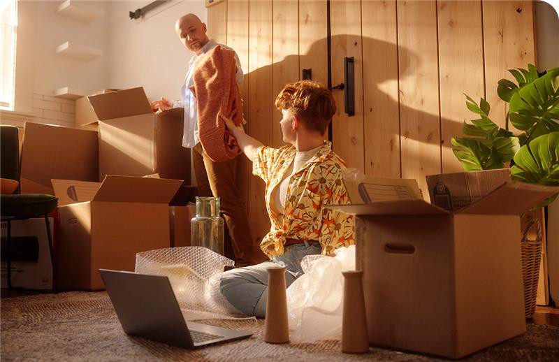 Two people unpacking boxes in a sunlit room, one sitting on the floor with a laptop while the other holds a sweater, surrounded by moving boxes.
