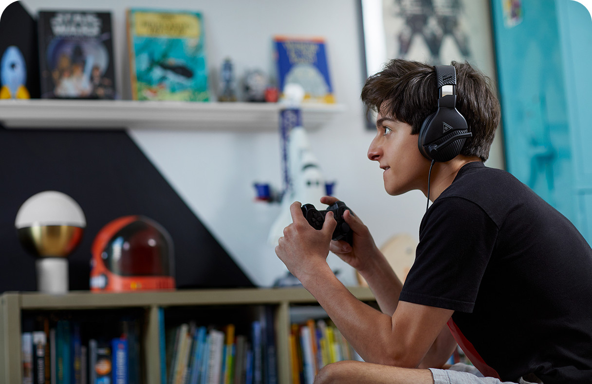 Boy wearing headphones playing a video game with a controller, sitting in a room with books and toys on shelves in the background.